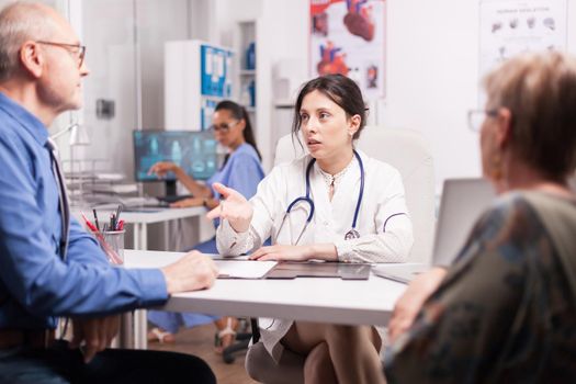 Female doctor explaining diagnosis to senior man elderly woman in hospital office during medical check up. Sick married pensioners. Nurse working on computer.