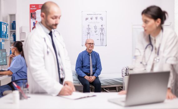 Two specialists doctors discussing senior patient diagnosis in hospital office. Scared mature man waiting medic expertise for his illness. Nurse working on laptop.