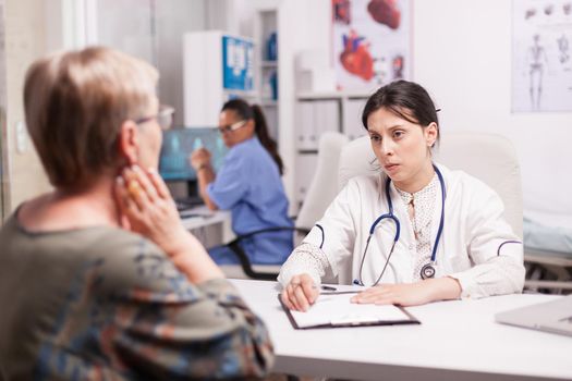 Young doctor examining senior patient in hospital cabinet wearing white coat and stethoscope. Mature woman with sore neck. Nurse working on computer.