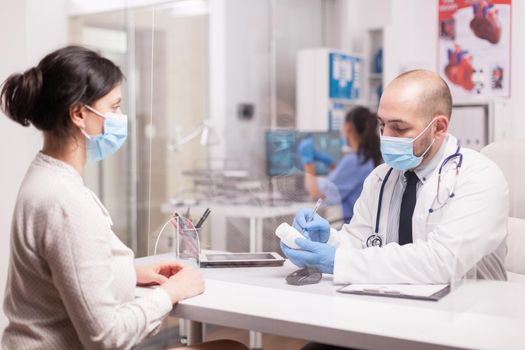 Doctor with face mask against coronavirus writing prescription on bottle pills for female patient in hospital office. Nurse in blue uniform working on computer.