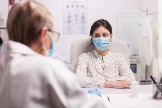 Portrait of worried patient with face mask against coronavirus during consultation with senior doctor in hospital office.