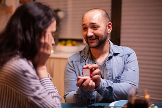 Young man asking wife to marry him during dinner in kitchen. Man making proposal to his girlfriend in the kitchen during romantic dinner. Happy caucasian woman smiling being speechless