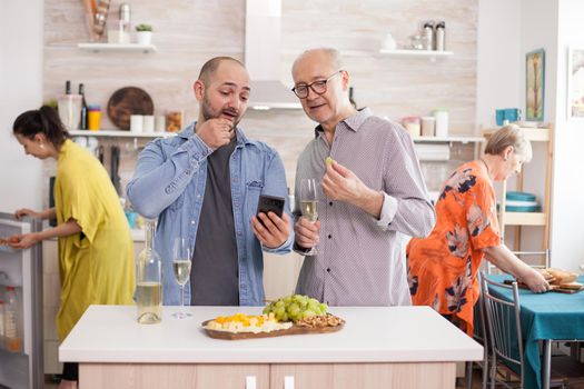Father and son watching video on smartphone during family brunch. Senior man holding a glass of white wine. Wife taking egg from refrigerator.