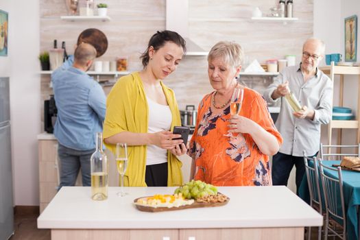 Mother and daughter watching video on smartphone in kitchen while enjoying a glass of wine. Senior father holding bottle of wine.