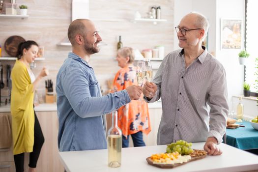 Happy son and father clinking wine glasses in kitchen during lunch with family.