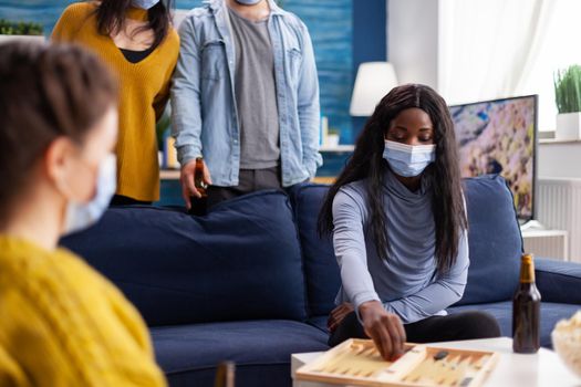 African woman competing with friends playing backgammon wearing face mask keeping social distancing during social pandemic with covid19 drinking beer in home living room. Board games, recreation, players. Conceptual image.