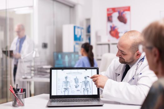 Doctor pointing at human skeleton on laptop in hospital office during consultation of old woman before surgery. Senior medic wearing white coat taking notes on clipboard in clinic corridor.