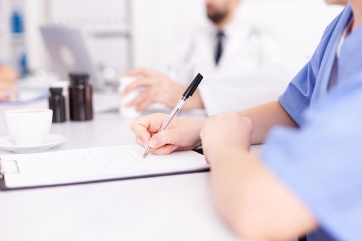 Medical woman practitoner writing notes on clipboard wearing blue uniform during conference with coworkers. Clinic expert therapist talking with colleagues about disease, medicine professional.