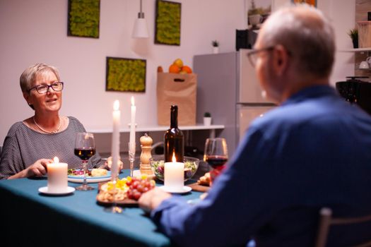 Mature woman having a conversation with husband while holding glass of red wine in kitchen. Senior couple sitting at the table in kitchen, talking, enjoying the meal, celebrating their anniversary in the dining room.