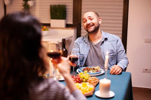 Husband feeling happy smiling at wife during romantic dinner. Talking happy sitting at table dining room, enjoying the meal at home having romantic time at candle lights.