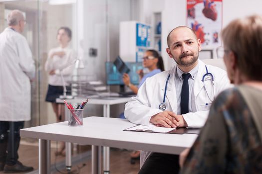 Young doctor looking worried at elderly aged patient during medical examination in hospital office. Young sick woman talking with mature medic on clinic corridor about treatment for her illness and nurse is holding x-ray.