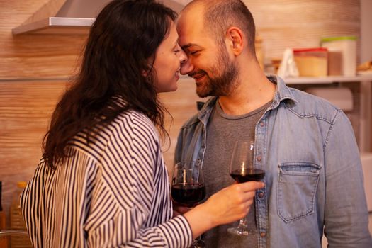 Couple keeping eyes closed during romantic date in kitchen. Adult couple at home, drinking red wine, talking, smiling, enjoying the meal in dining room.