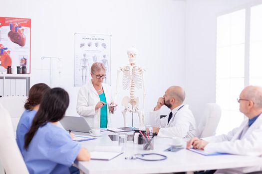 Mature surgeon doing demonstration on human anatomy on skeleton for medical staff in conference room. Clinic expert therapist talking with colleagues about disease, medicine professional