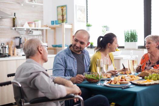 Son looking at disabled senior father in wheelchair during family lunch with healthy food. Happy Mature parents.