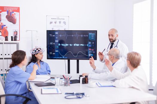 Doctor discussing about braing waves while is wearing headset with sensors. Monitor shows modern brain study while team of scientist adjusts the device.