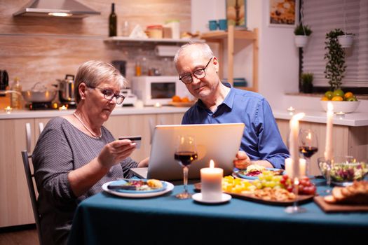 Old woman paying online using credit card and laptop in kitchen. Old people sitting at the table, browsing, using the technology, internet, celebrating their anniversary.