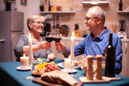 Elderly couple enjoying a glass of wine during festive relationship celebration. Happy cheerful senior elderly couple dining together in the cozy kitchen, enjoying the meal.