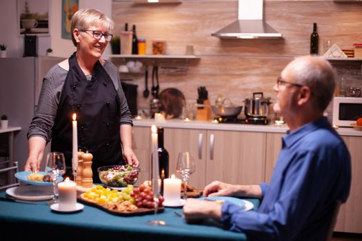 Husband sitting at dinner table and wife serving tasty food for romantic relationship anniversary. Elderly old couple talking, sitting at the table in kitchen, enjoying the meal,