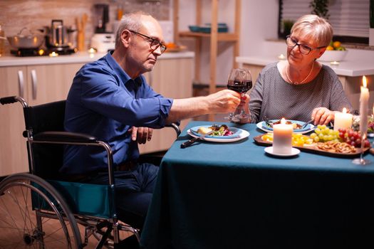 Man with disabilities having dinner with wife and clinking wine glass. Wheelchair immobilized paralyzed handicapped man dining with wife at home, enjoying the meal