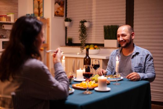Romantic dinner with man in foreground feeling happy with wife in dining room. Relax happy people clinking, sitting at table in kitchen, enjoying the meal, celebrating anniversary.