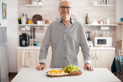 Cheerful mature man in kitchen smiling at camera with assorted cheese and grapes.