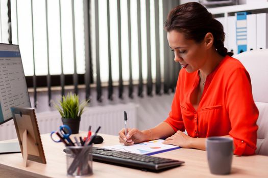 Businesswwoman entrepreneur writing financial sales on charts sitting at desk in workplace wearing red. Successful financial auditor in data marteking looking at graphs on computer screen and writing report.