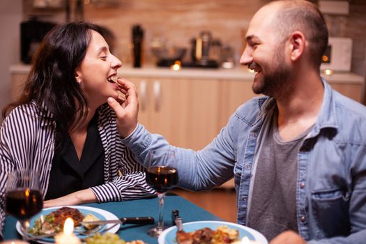 Husband feeding wife with grapes during romantic dinner celebrating relationship. Wife and husband celebrating anniversary with red wine, tender moments at candle lights in kitchen.