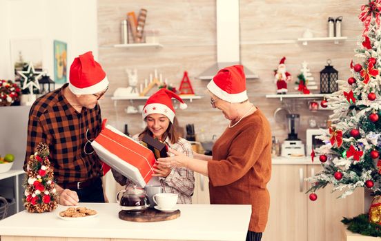 Smiling child during christmas celebration receiving gift box from cheerful senior man and woman. Happy loving grandparents celebrating winter holidays and relationship with granddaughter in home with x-mas decoration.