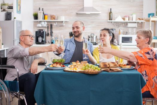 Multi generation family toasting glasses. Disabled senior man in wheelchair clinking wine glass with son , daughter and mature wife.