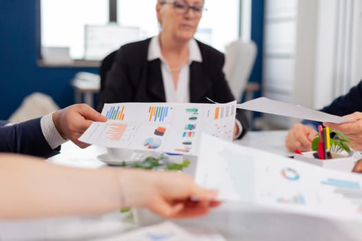 Confident company manager giving working tasks to diverse teamworkers analysing paperwork with graphs sitting in start up office. Colleagues working in office at conference table in startup management company.