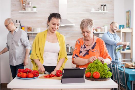 Mother and daughter following online recipe on tablet for healthy salad. Husband smiling while holding bottle of wine. Senior man opening refrigerator door in kitchen.