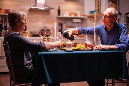 Senior man opening wine bottle during romantic date with wife. Elderly couple sitting at the table in kitchen, talking, enjoying the meal, celebrating their anniversary in the dining room.