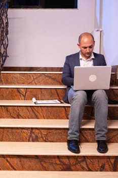 Overworked tired businessman at workplace working on deadline for job project using notebook. Confident corporate entrepreneur using laptop doing overtime sitting on stairwell.