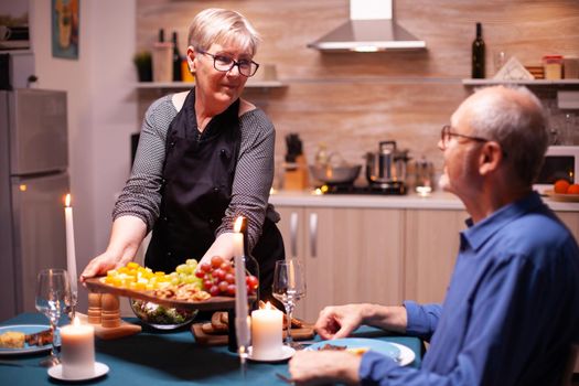 Senior man and woman enjoying romantic dinner in kitchen. Elderly old couple talking, sitting at the table in kitchen, enjoying the meal, celebrating their anniversary .