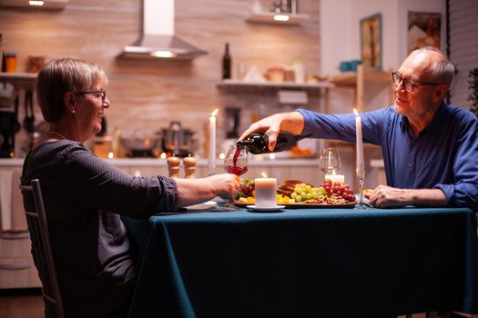 Senior man serving wife with wine during romantic dinner. Elderly couple sitting at the table in kitchen, talking, enjoying the meal, celebrating their anniversary in the dining room.