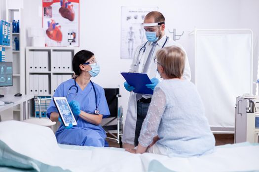 Nurse consulting with doctor during examination of senior woman in hospital room holding tablet pc and wearing face mask against covid-19 as safety precaution. Medical control for infections,