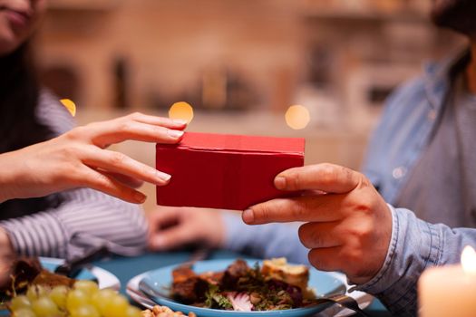Close up of gift box holded by husband and wife during romantic dinner. Cheerful man dining with woman at home, enjoying the meal, celebrating their anniversary at candle lights