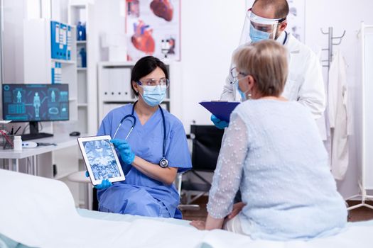 Medical staff explaining diagnosis to senior woman in hospital room using tablet pc and wearing face mask against coronavirus as safety precaution. examination for infections, disease and diagnosis.