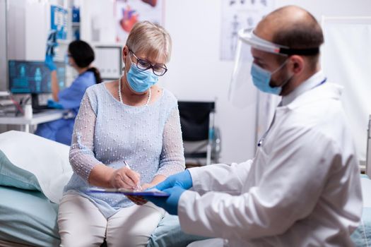 Elderly patient signs medical agreement of test results wearing protective mask against covid pandemic in hospital. Healthcare medical physician consultation during COVID-19 global crisis.