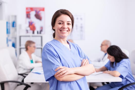 Young nurse wearing medical uniform in hospital conference room smiling at camera. Friendly medical practitioner in clinic meeting room, robe, specialist.