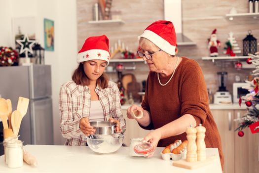 Granddaughter and mother on christmas day doing cookies with love. Happy cheerful joyfull teenage girl helping senior woman preparing sweet biscuits to celebrate winter holidays wearing santa hat.