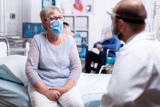 Elderly woman discussing with doctor during consultation in hospital examination room wearing face mask agasint covid-19. Global health crisis, medical system during pandemic, sick elderly patient.