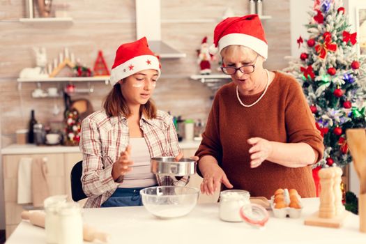 Child on christmas day with xmas tree in the background baking traditional cookies. Happy cheerful joyfull teenage girl helping senior woman preparing sweet biscuits to celebrate winter holidays wearing santa hat.