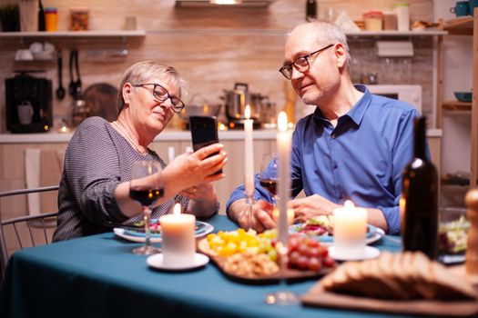 Old couple using phones in the kitchen during romantic dinner. Sitting at the table in the dining room , browsing, searching, using phone, internet, celebrating their anniversary in the dining room.