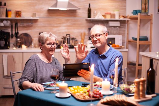 Old husband and wife waving at tablet pc while celebrating relationship with festive dinner. Couple sitting at the table, browsing, talking, using internet, celebrating their anniversary in dining room.