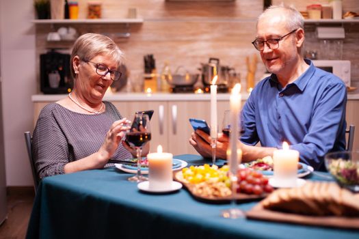 Elderly couple talking and using phone sitting at table with candles during dinner . Sitting at the table in the kitchen, browsing, searching, internet, celebrating their anniversary in the dining room.