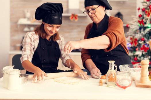 Granddaughter in apron preparing cake on christmas day with grandmother. Happy cheerful joyfull teenage girl helping senior woman preparing sweet cookies to celebrate winter holidays.