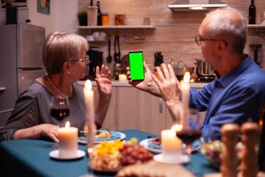 Elderly man and woman waving at phone with green screen. Aged people looking at mockup template chroma key isolated smart phone display using techology internet sitting at the table in kitchen.