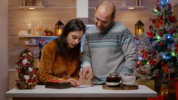 Cheerful couple preparing gift and christmas card to give present box to relatives for holiday festivity. Festive man and woman signing postcard on christmas eve for seasonal celebration.