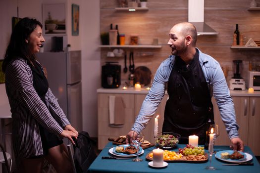 Cheerful husband surprising wife with dinner in dining room after she arrives from work. Man preparing festive dinner with healthy food, cooking for his woman a romantic dinner,